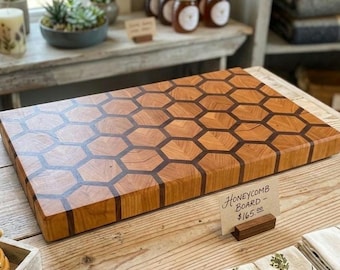 May include: A rectangular wooden cutting board featuring a geometric honeycomb pattern created with light and dark wood segments. A small sign in front of the board displays the handwritten text Honeycomb Board - $165.00 on a light tan card.