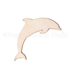 May include: A light brown wooden dolphin cutout against a white backdrop. The dolphin is shown leaping, with its body arched and tail up. The cutout has a smooth finish and seems to be made from thin wood. The dolphin is a popular sea creature.