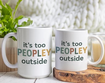 May include: Two white coffee mugs with the text "it's too PEOPLEY outside" in colorful letters. The mugs are sitting on a wooden surface with a white blanket in the background.