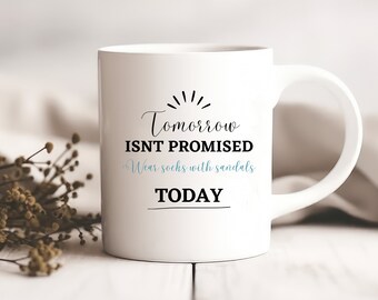May include: White ceramic mug with black text that reads "Tomorrow isn't promised. Wear socks with sandals today."