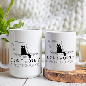 Puede incluir: Dos tazas blancas con asas negras. Cada taza tiene un gato negro sentado en un portátil con el texto "No te preocupes, soy del soporte técnico".