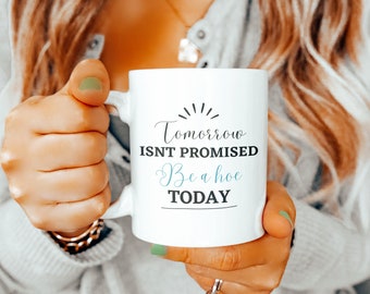 May include: White ceramic mug with the text "Tomorrow ISNT PROMISED Be a hoe TODAY" in black and teal script. The mug has a handle and is held by a person.