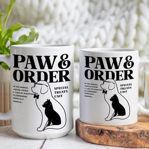 May include: Two white ceramic mugs with black text and illustrations. The mugs feature the text "PAW & ORDER" and a dog and cat wearing bow ties. The text below the illustrations reads "In the criminal justice system, a lack of scratches is considered especially heinous..." and "Special Treats Unit".