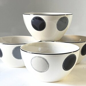 Peut inclure: Un ensemble de quatre bols en céramique blanc cassé avec un bord noir et un motif de grands points circulaires dans des tons de gris foncé et gris clair. Les bols sont disposés en groupe, mettant en valeur leurs surfaces extérieures lisses.