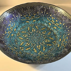 May include: A round glass bowl featuring an intricate, embossed mandala-style pattern in shades of iridescent blue, teal, and purple. The surface has a textured, raised relief design that radiates from the centre towards the scalloped rim.