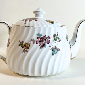 May include: White ceramic teapot with a lid, handle, and spout. The teapot features a swirling design and is decorated with floral patterns in shades of pink, yellow, and green. Gold trim accents the edges.