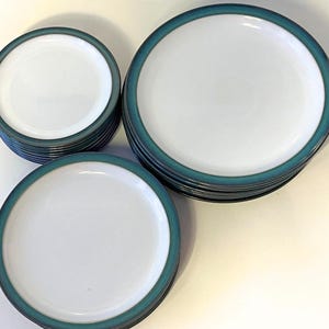 Puede incluir: Un conjunto de platos redondos de cerámica con un centro blanco y un borde ancho de color verde azulado oscuro. El juego incluye varios platos pequeños apilados y una pila de platos más grandes, todos con un acabado liso y brillante.