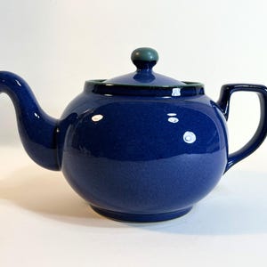 May include: A navy blue ceramic teapot with a rounded body, curved spout, and a square handle. The lid has a green knob. The teapot is a classic design for brewing tea.