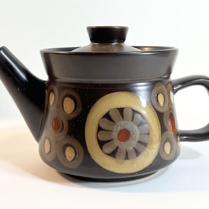 Puede incluir: Una tetera de cerámica marrón con tapa, boquilla y asa. La tetera presenta un patrón decorativo de círculos y diseños florales en tonos marrones, beige y naranja. La tetera está diseñada para preparar té.