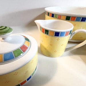 May include: A ceramic sugar bowl with a lid, a creamer jug, and a large bowl. Each piece features a yellow base with a small orange diamond pattern, a colourful border of red, green, blue, and yellow squares, and a white interior finish.
