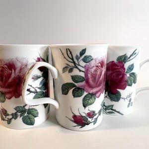 May include: Three white ceramic mugs featuring a botanical pattern of deep pink and red roses with green leaves and thorny stems. Each mug has a smooth cylindrical body and a curved handle, displaying detailed floral illustrations on the exterior.