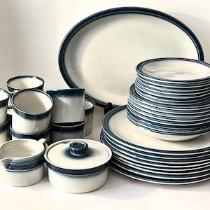Peut inclure: Un service de vaisselle en céramique composé d'une base couleur crème avec des bords bleu foncé. L'ensemble comprend un grand plat ovale, plusieurs assiettes empilées, des bols, des tasses, un petit pot à lait et un bol avec couvercle.