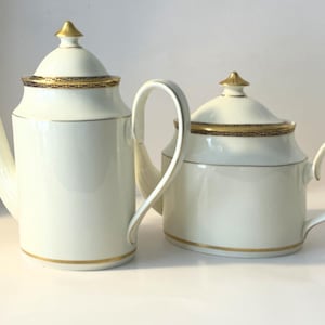 May include: A tall coffee pot and a shorter teapot, both crafted from off-white ceramic. Each piece features a matching lid with a gold-toned finial, a gold-trimmed rim with a dark patterned band, and a thin gold line around the base.