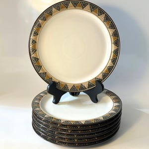 May include: A stack of seven round ceramic plates with a cream-colored center and a wide border featuring a repeating geometric pattern of black, gold, and white triangles. One plate is displayed upright on a black plastic stand for visibility.