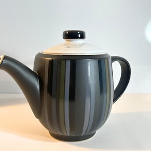May include: A black and white ceramic teapot with vertical stripes in shades of grey and green. The teapot has a curved spout, a rounded body, and a black handle. The lid is white with a black knob.