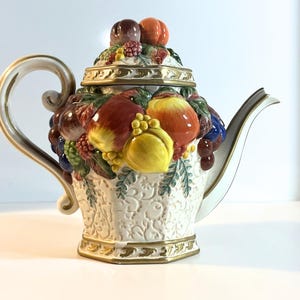 May include: A decorative ceramic teapot with a fruit basket design. The teapot has a cream-coloured base with embossed patterns and is adorned with colourful, three-dimensional fruits, including apples, grapes, and lemons. The handle and spout are white with gold trim.