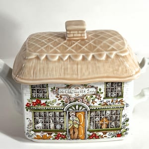 May include: A ceramic teapot shaped like a cottage with a tan thatched roof lid and a chimney. The white body features a colorful illustration of a building labeled Ye Olde Tea Pot Inn, complete with windows, flowers, and a person in the doorway.