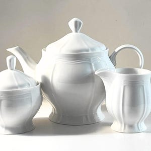 Peut inclure: Un service à thé en céramique blanche composé d'une théière avec couvercle, d'un sucrier avec couvercle et d'un pot à lait. Chaque pièce présente une finition lisse et brillante avec des panneaux cannelés verticaux et une bande horizontale.