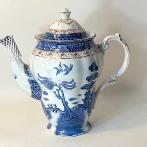 May include: A white porcelain teapot featuring a blue willow pattern with a matching lid. The design includes depictions of birds, trees, and architectural structures. The teapot has a curved handle, a spout with a lattice pattern, and gold trim.
