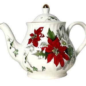 May include: A white ceramic teapot featuring a festive botanical pattern of vibrant red poinsettia flowers, green ivy leaves, and white hellebore blossoms. The teapot has a fluted body, a curved handle, a spout, and a lid topped with a gold-coloured knob.