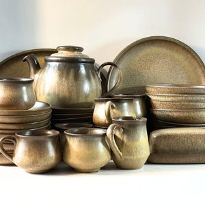 Puede incluir: Un juego de vajilla de cerámica que incluye una tetera, tazas, platos, cuencos y una mantequera. Cada pieza tiene un esmalte moteado en tonos tierra marrón y bronceado con un patrón de goteo más oscuro en los bordes, creando un aspecto uniforme.