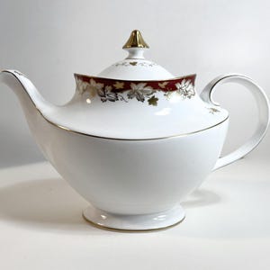 Peut inclure: Théière en porcelaine blanche avec bordure et accents dorés. Le couvercle et le bord présentent une bande bordeaux avec des motifs de feuilles d'or. La poignée est incurvée et le bec est fin. La théière repose sur une petite base ronde.