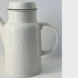 May include: A white ceramic coffee pot featuring a raised floral relief pattern on the body. The pot includes a matching lid with a thin black rim, a curved handle on the right side, and a tapered spout on the left side, all in a glossy finish.