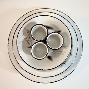 Peut inclure: Un ensemble de trois assiettes en céramique empilées et trois petites tasses assorties disposées au centre. La vaisselle présente un émail moucheté blanc cassé avec des bords irréguliers brun foncé et des motifs botaniques noirs.