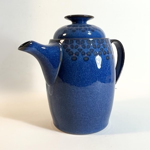 Puede incluir: Una cafetera de cerámica azul cobalto con tapa y asa. La cafetera tiene un patrón moteado en la parte superior y un acabado brillante. El pico es curvo y el asa es de color azul oscuro sólido.