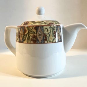 May include: A white ceramic teapot featuring a decorative band around the upper portion. The band displays a pattern of red flowers, green leaves, and geometric diamond shapes in shades of blue and tan. The teapot includes a matching lid with a knob.