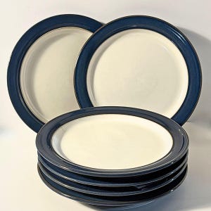 May include: A set of round ceramic plates featuring a cream-colored center and a wide, dark navy blue rim. The collection includes two plates standing upright in the back and a stack of four identical plates positioned in the foreground.