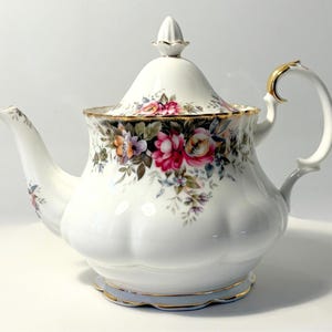 May include: A white porcelain teapot featuring a scalloped base and a floral pattern with pink, red, and orange roses. The teapot includes a matching lid with a pointed finial, a curved handle with gold trim, and gold accents along the rim and base.