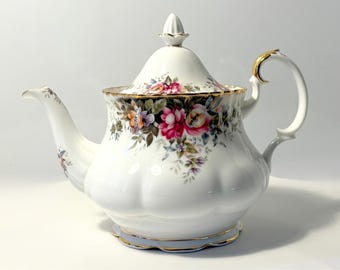 Pode incluir: Uma bule de porcelana branca com base festonada e um padrão floral com rosas cor-de-rosa, vermelhas e cor de laranja. O bule inclui uma tampa a condizer com um remate pontiagudo, uma pega curva com rebordo dourado e detalhes dourados.