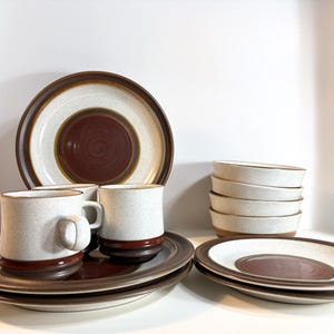 May include: A stoneware dinnerware set featuring a speckled off-white glaze with dark brown circular accents. The collection includes two mugs, several flat plates, and a stack of four small bowls, all sharing a consistent earthy colour palette.
