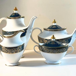 Peut inclure: Un service à thé en porcelaine de quatre pièces comprenant une grande théière, une théière plus basse, un pot à lait et un sucrier avec couvercle. Chaque pièce est blanche avec une bande bleu foncé ornée de motifs floraux dorés.