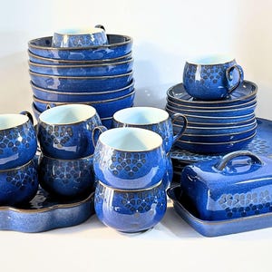 Puede incluir: Un juego de vajilla de cerámica azul con un patrón floral azul oscuro repetitivo. El conjunto incluye varias tazas redondeadas, platillos apilados, cuencos, una mantequera con tapa y una bandeja de servir festoneada, todo con acabado brillante.