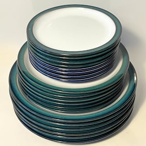 May include: A stacked set of round ceramic plates featuring a white centre and a wide, dark teal rim. The collection consists of multiple plates of varying diameters, arranged in three distinct tiers from smallest to largest, creating a uniform look.