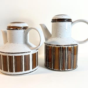 May include: Two ceramic coffee pots with white and brown vertical stripes. The pots have a white body, brown lids, and large handles. The design is a mix of modern and vintage styles. The pots are likely for serving coffee or tea.