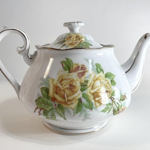 May include: A white porcelain teapot with a lid, decorated with yellow rose floral patterns and gold trim. The teapot has a curved handle and spout. A classic piece for serving tea, perfect for afternoon tea parties.