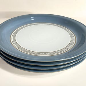 May include: A stack of four round ceramic plates featuring a solid dusty blue rim and a pale white centre. A decorative band with a repeating circular pattern in dark grey and white separates the blue rim from the white centre of each plate.