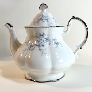 May include: A white porcelain teapot with a silver trim and handle. The teapot features a floral pattern in shades of blue and gray. The lid is topped with a decorative finial. The teapot is designed for serving tea.