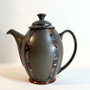 Może przedstawiać: Nakrapiany szary ceramiczny imbryk z pokrywką i uchwytem. Czajnik posiada ozdobny pas geometrycznych kształtów w odcieniach brązu, błękitu i bieli. Podstawa i gałka pokrywy są ciemnobrązowe.
