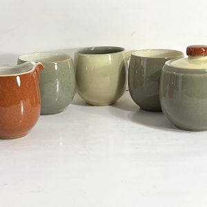 Peut inclure: Un ensemble de cinq récipients en céramique comprenant un pot à lait couleur rouille avec un bec verseur, trois tasses arrondies dans des tons variés de vert sauge et crème, et un sucrier vert sauge avec un couvercle crème et un bouton rouille.