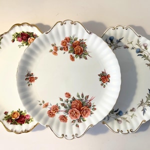 Puede incluir: Tres platos de cerámica blanca con bordes festoneados y ribete dorado. El plato izquierdo presenta racimos de rosas rojas y amarillas. El plato central muestra ramos de rosas naranjas. El plato derecho exhibe motivos florales de cardos azules.