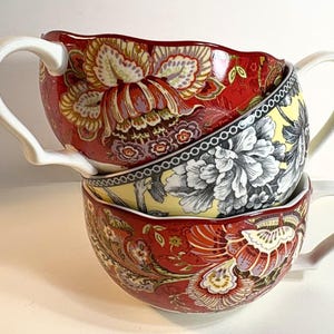 Puede incluir: Dos tazas de cerámica apiladas con bordes festoneados y asas blancas. La taza superior presenta un fondo rojo con un gran patrón floral en amarillo, blanco y morado. La taza inferior muestra un fondo amarillo con diseños florales en blanco y negro.