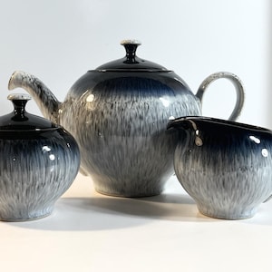 Puede incluir: Un juego de té de cerámica de tres piezas con un esmalte moteado azul oscuro y gris. El juego incluye una tetera con tapa, un azucarero con tapa y una jarra de crema. La tetera tiene un pico y un asa curvados.