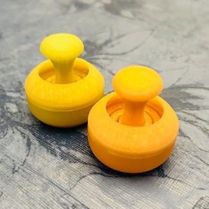 Może przedstawiać: Dwa plastikowe wykrawacze do ciasteczek w kształcie grzybów, jeden żółty, a drugi pomarańczowy. Wykrawacze znajdują się na teksturowanej powierzchni.