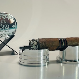 Könnte beinhalten: Eine Sammlung silberner Zigarrenaccessoires, darunter ein Aschenbecher, eine Zigarrenablage und Untersetzer. Ein Untersetzer zeigt den Text "NO. 82" mit dekorativen Elementen. Eine brennende Zigarre liegt auf einem silbernen Ständer.