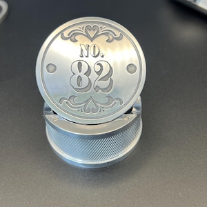 Könnte beinhalten: Ein silberner Metallzerkleinerer mit rundem Deckel mit der Nummer "82" und dekorativen Verzierungen. Der Sockel hat ein strukturiertes, gebogenes Design. Der Zerkleinerer wird wahrscheinlich für Kräuter oder Gewürze verwendet.