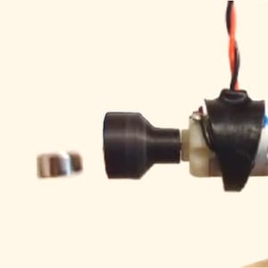 May include: A small black electric motor with two black plastic end caps, suspended by wires. Two silver metal discs are visible on either side of the motor.